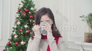 快乐的年轻亚洲女人，在圣诞节的时候，在家里的客厅里，抱着温暖的咖啡，微笑着对着镜头。